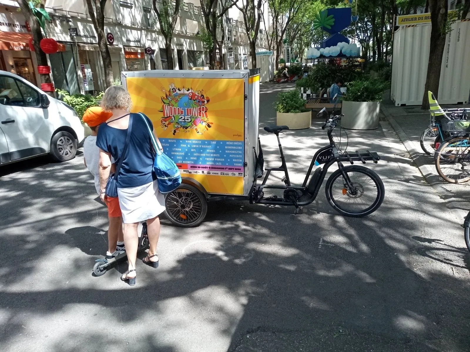 Triporteur publicitaire LCDG habillé aux couleurs d'un festival dans une rue arborée animée